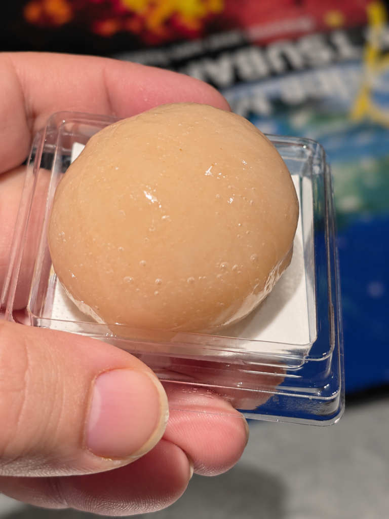 Une main tient une bouchée ronde et beige dans une petite barquette transparente.