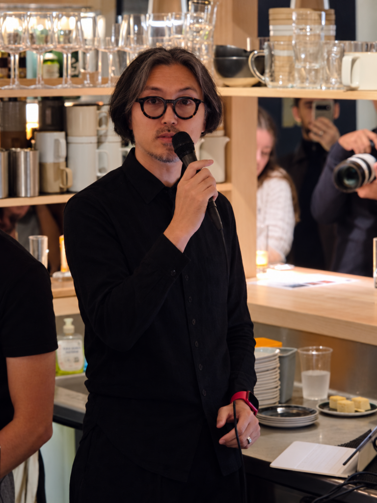 Takao Mishima, directeur général délégué de l'entreprise familiale japonaise Mishima Foods, vêtue de noir et portant des lunettes s’exprime au micro devant les étagères de bouteilles du bar.