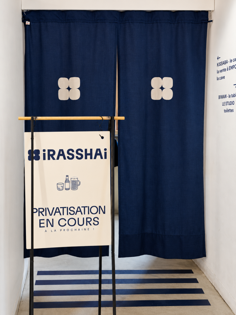 Un rideau japonais bleu à motifs blancs marque l’entrée d’un espace, devant un panneau Irasshai indiquant « Privatisation en cours ».