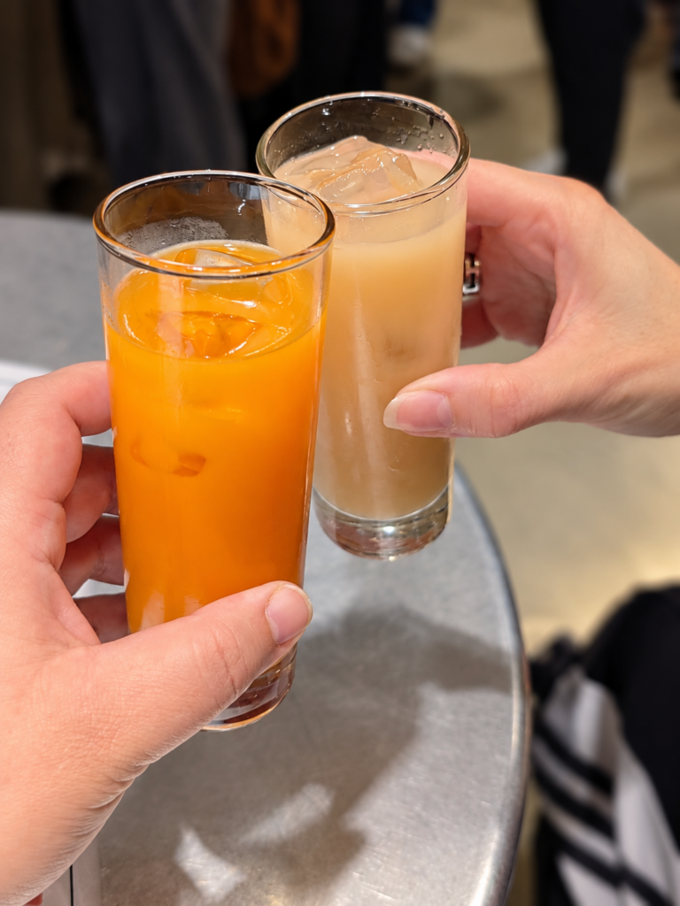 Deux mains rapprochent de grands verres de cocktails avec des glaçons, l’un orange et l’autre rose pâle.