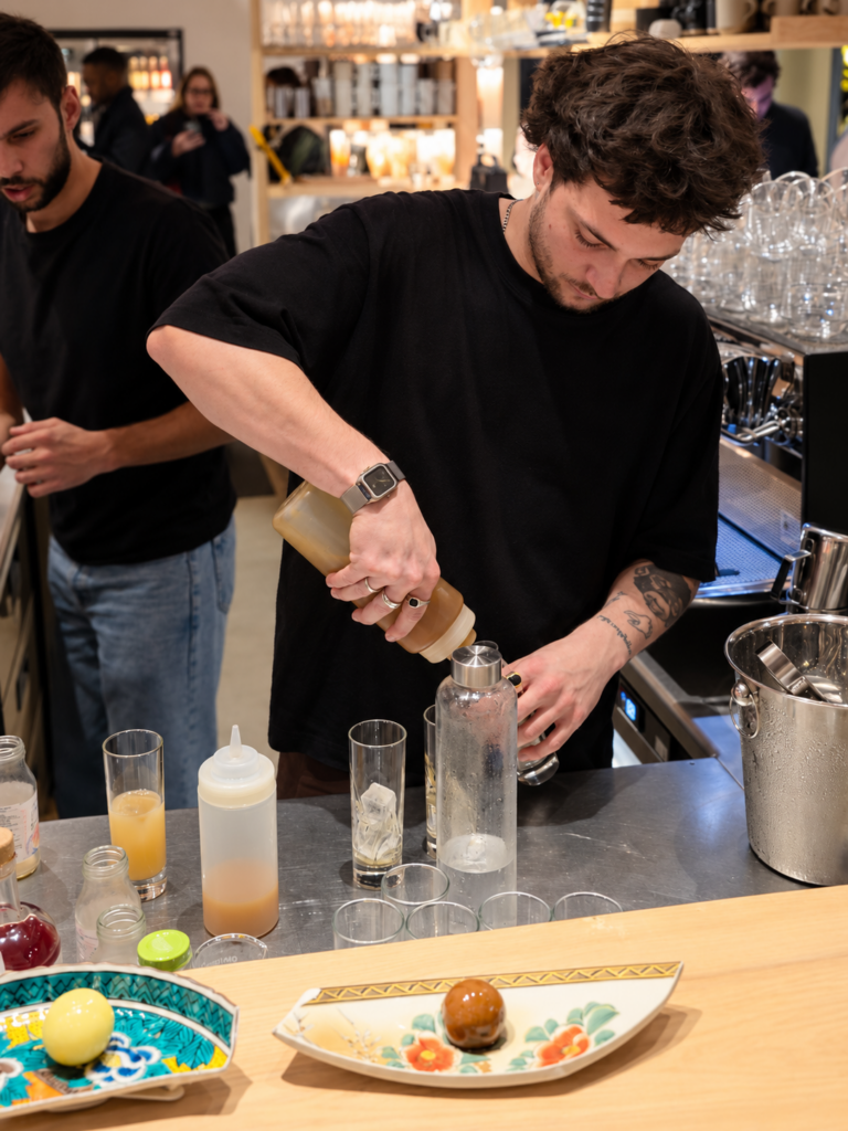 Un barman verse une préparation dans un verre derrière le comptoir, au milieu des bouteilles et des ustensiles de bar.